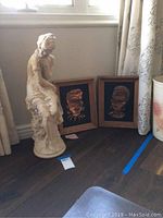 Full view of the Greco-Roman lady statue seated next to two vintage copper framed etched wall art pieces depicting faces with ethnic motifs