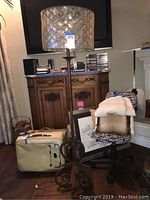Full view of the floor lamp standing near a wooden cabinet and a chair. The metal base features scroll work and the shade is composed of translucent Capiz shell segments in a honeycomb pattern.