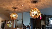 Pair of spherical brushed gold metal chandeliers hanging from ceiling, showing detailed scrollwork casting shadows on ceiling.