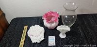 Four glass pieces displayed on black cloth: small white milk glass hobnail vase, white and pink ruffled glass vase, white open hand-shaped dish with floral and butterfly decoration, clear glass vase with flared rim.