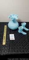 Blue Fenton glass boots and blue vase displayed on black tablecloth with measuring tape for scale