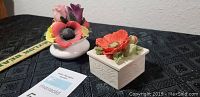 Two ceramic trinket boxes on black cloth background, one round with colorful flower bouquet lid, one square with single large flower lid