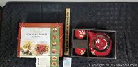 Red ceramic teapot with two matching tea cups by California Pantry and boxed Numi Flowering Tea Set with glass teapot and tea on black textile background with wood scale for size.