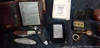 Display of brown leather wallet, two folding pocket knives, Zippo lighter with original inserts, a black box, and small vintage cigarette case with tobacco-related items.