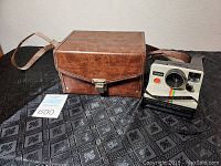 Front view of the Polaroid Land Camera OneStep next to its brown leatherette carrying case with shoulder strap on a black patterned surface.