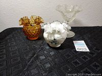 Three glass ripple edge pieces on a black patterned tablecloth: amber hobnail vase, white milk glass bowl with blue floral design, clear pedestal bowl with ruffled edge.