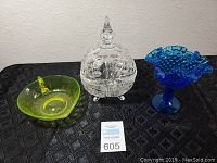Front view of three pieces on black tablecloth: yellow-green glass dish, clear cut crystal candy dish with lid, and blue hobnail ruffled compote.