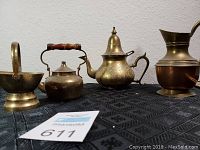 Photo showing five brass decor items on a black patterned cloth: pedestal bowl, two teapots, pitcher and sugar bowl.