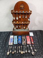 Full view of wooden spoon rack with 12 spoons hanging and 7 spoons laid out on table, including some in packaging.