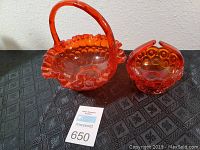 Both orange glass baskets shown side by side on black patterned cloth with auction label and ruler nearby for size reference