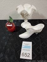 Three decorative glass items on black cloth: a red glass apple figurine with green leaf, a white hobnail milk glass shoe, and a white hobnail milk glass compote with ruffled edge.