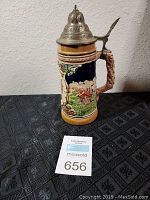 Ceramic German beer stein with hinged pewter lid and detailed rural scene, placed on black patterned surface, with printed auction tag number 656 in front.