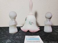 Photo of pink frosted glass bell with floral decoration and two frosted glass praying child figurines on black patterned cloth.