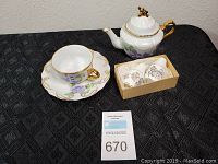 Photo of the full set including teapot with lid, tea cup and saucer, and boxed salt and pepper shakers on a black patterned cloth.