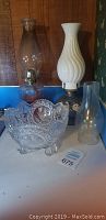 Three items displayed together on a white surface against wood and wallpapered walls: a clear glass oil lamp with chimney, an oil lamp with white swirled glass shade and decorated base, and a clear lead crystal dish with intricate cut patterns.