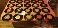 Photo showing a flat lay array of 26 vintage 45 RPM vinyl records on a wooden table, various labels visible including orange, yellow and green ones