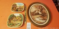 Three art pieces shown together on table; two ceramic plates with cottage scenes and one oval framed swan artwork.