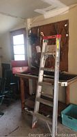 Image showing aluminum 6ft Werner stepladder standing in garage next to wooden workbench and pegboard with tools.