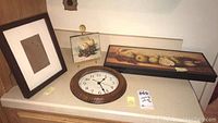 Overview of the 12-inch Ingraham round wall clock, a photo frame, and a long rectangular framed fruit print wall art on a countertop.