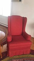 Red wing back chair shown from front left side near window and table, upholstered in red fabric with pleated skirt and padded arms.