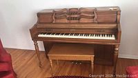Full view of Wurlitzer upright piano with bench in wooden finish on hardwood floor against white wall