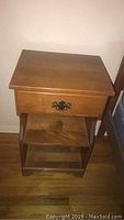 Wooden side table with one drawer, two shelves, metallic drawer pull, sits on hardwood floor, note wear and dings on surfaces.