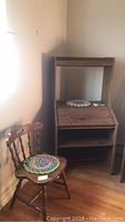 Photo showing wood desk with slant front, adjustable top shelf, and two lower open shelves, with handwoven doily on top, next to wood chair with handwoven pad.
