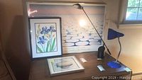 Photo of two desk lamps on a dark wood table along with three framed artworks leaning against the wall.