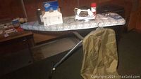 Photo showing ironing board with various items on and around it including iron, Scotchguard spray, clothing bag, and detergent.