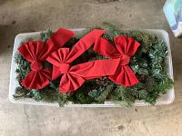 Plastic storage bin filled with green artificial pine garland with natural-style pinecones and three large red bows on top.