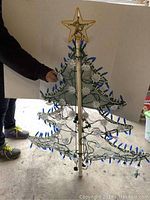 Green wire-frame Christmas tree lit with blue bulbs, gold star topper, and white snowflake decorations shown upright with person holding it for scale.
