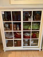 Front view of the white wooden display cabinet with sliding glass doors and grid pattern, filled with various household items.