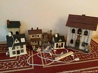 Photo showing six Americana decorative houses in various sizes and shapes on a red patterned rug, some with power cords attached and a wooden figurine in front.