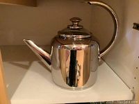 Polished stainless steel tea kettle with lid, curved spout and knob on lid, placed on shelf