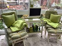 Two white wooden garden chairs with matching green patterned cushions and green pillows plus blankets. Small white table with glass top, decorative figurines on lower shelf, and Toshiba flat screen TV on top. Window background.