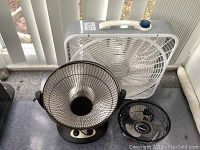 White box fan heater with handle, blue dial on top, black standing heater with circular heating element and two control knobs on base, small black Honeywell desk fan with grille and three blades. Items positioned on a floor next to window blinds.