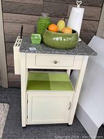 Front view of cream colored rolling cart with granite look chopping block top, drawer, green bowl with fake fruit, green jar, small green dish and cream colored paper towel holder