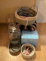 Heated dog beds stacked on top of each other alongside a pile of disposable blue pet pads, an automatic water dispenser with a clear plastic reservoir attached to a gray base with a feeding bowl, and a small beige dish with grooming items including a pet comb and packaging.