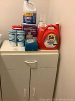 White storage cabinet with various cleaning supplies on top including Tide detergent, Clorox cleaner, Lysol wipes, and wastebasket liners box.