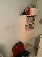 White bookshelf cabinet with DVDs, VCR tapes, red lamp, and white wicker container on top, mounted on wall