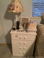 Photo of the white nightstand with four drawers, showing the floral lamp, stack of books, eyeglass holder, and various small items on top.