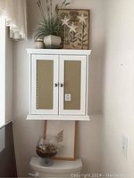 White wooden wall cabinet with two mirrored doors shown installed above toilet. Decorative items on top include faux grass in ceramic planter, star-shaped ornaments on sticks, small ceramic jar, framed botanical print.