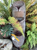 Photo of three-tier woven basket style outdoor fountain surrounded by garden plants, showing natural wear and leaves inside basins.