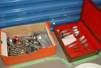 Box of assorted loose silver plated flatware and serving utensils next to a velvet-lined wooden flatware chest.