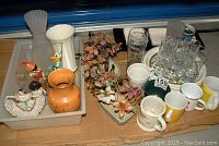Wide shot showing assorted ceramic and glassware items including figurines, vases, mugs, and decorative trees arranged in trays.