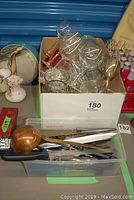 Photo showing box with clear glassware pieces including wine glasses, ramekins, and a silver-plated goblet, along with kitchen utensils and a ladle with wooden handle in a plastic bin.