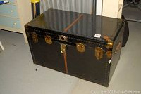 Front view of black McBrine Solidon trunk showing central latch, two side locks, riveted design with leather vertical strip on lid and front.