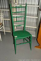 Full view of a green painted wooden antique high-back chair with five horizontal slats and four legs, standing on a concrete floor by metal shelves.
