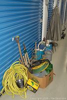 Photo showing various garden tools leaning against storage unit, yellow garden hoses coiled on floor, metal watering can, and lawn decor.