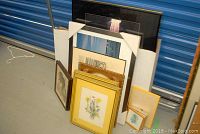 Photo of framed prints and photographs leaning against white foam in storage unit. Various frame colors and artwork visible.